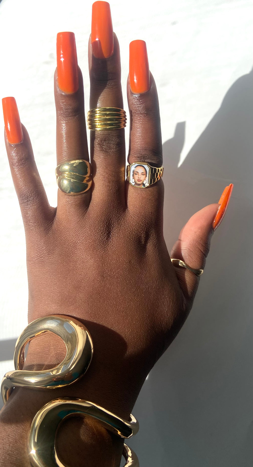 Hand wearing stacked gold rings and sculptural gold cuffs, including a square Sade-inspired promise ring, styled in natural sunlight.