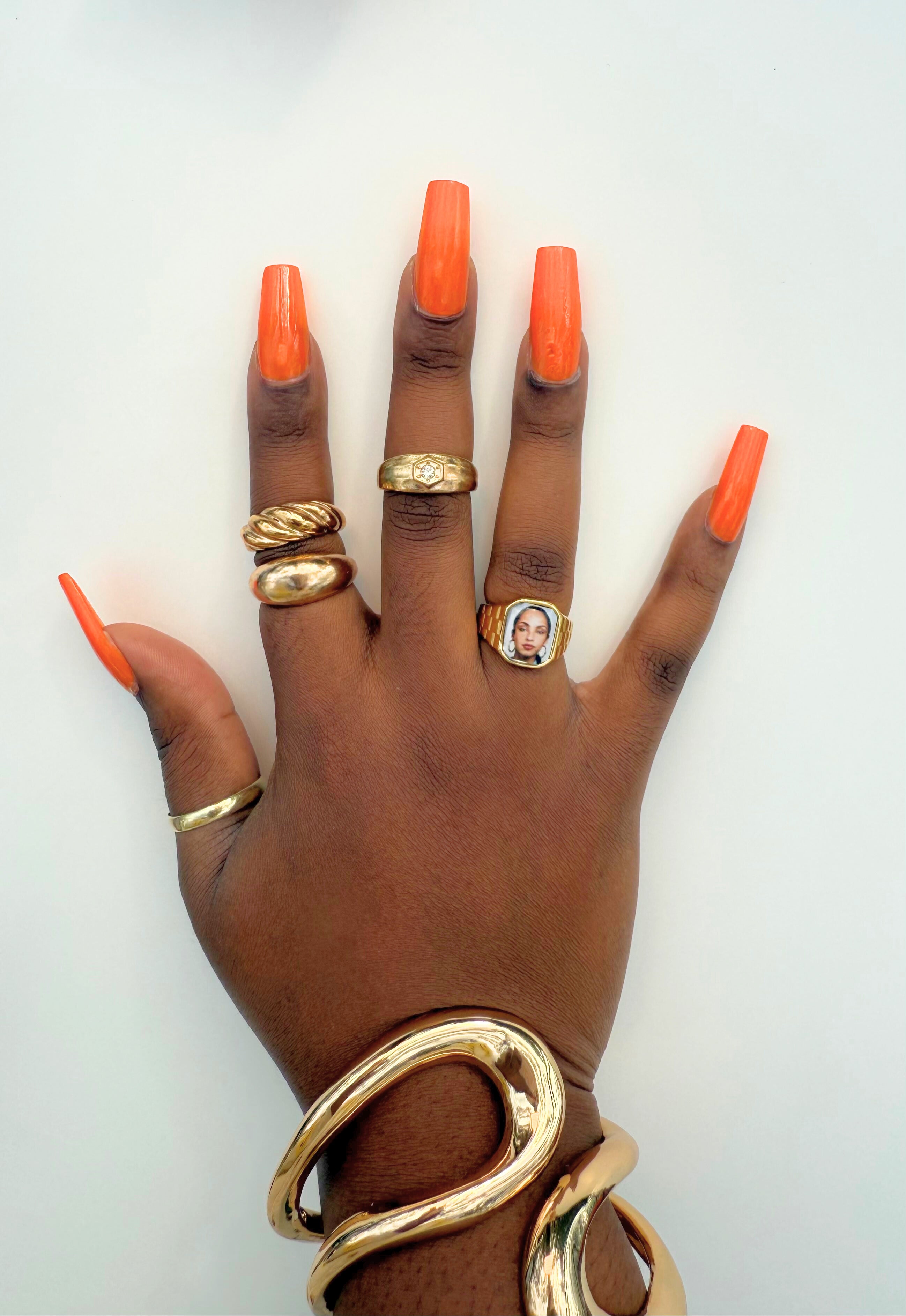 Hand wearing stacked gold rings and sculptural gold cuffs, including a square Sade-inspired promise ring, styled in natural sunlight.